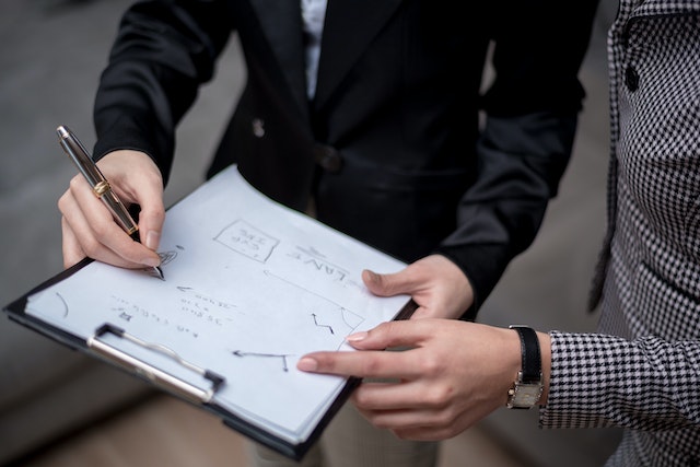 A Person in Black Suit Writing on paper on a clipboard while someone standing next to them is pointing at the paper